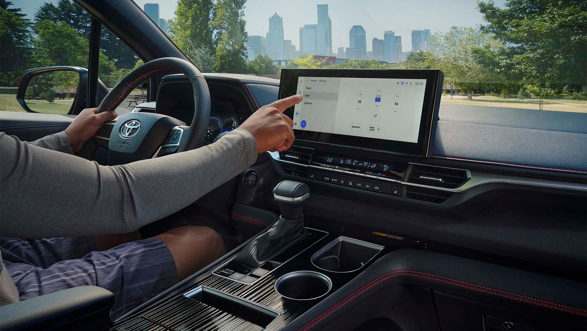 A person inside a 2025 toyota sienna is interacting with the large touchscreen display on the dashboard. The gear shift and cup holders are in view.