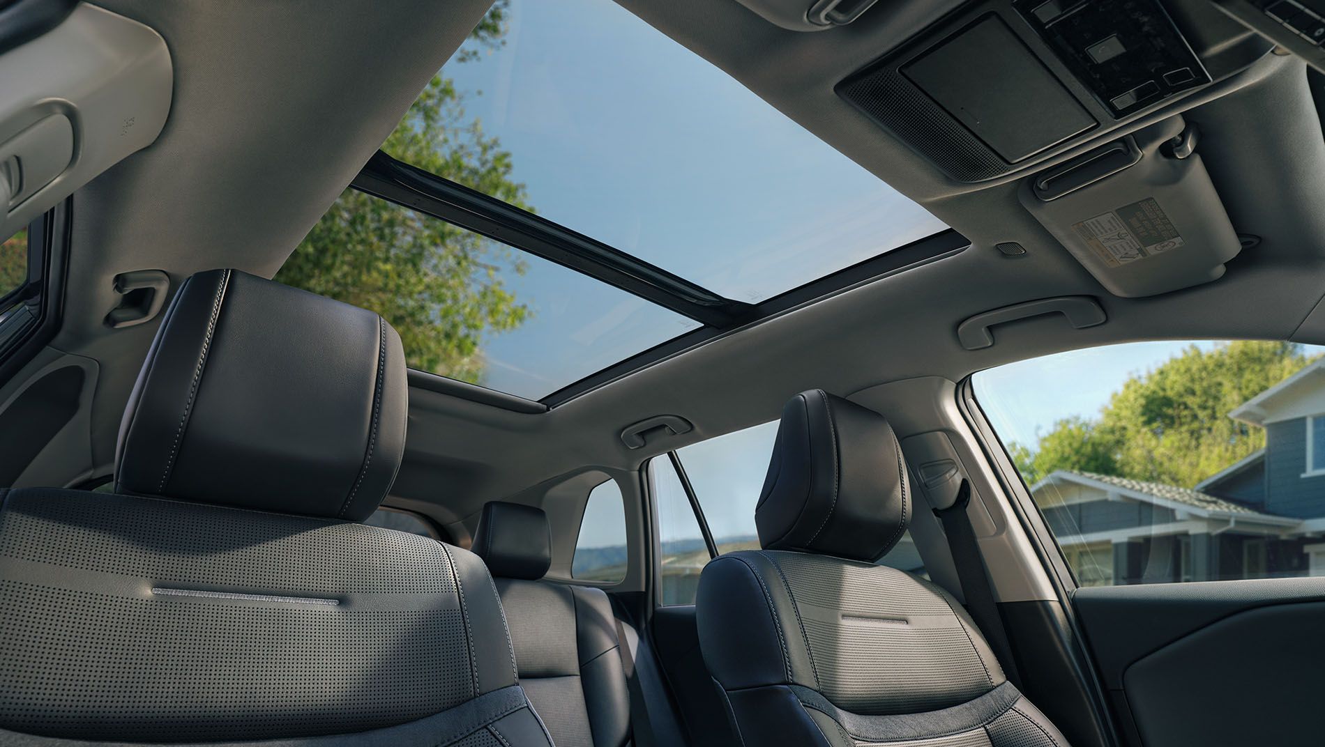 View from the back seat of a car interior, showing black leather front seats and a large panoramic sunroof with blue sky, trees, and part of a house visible through the glass roof.