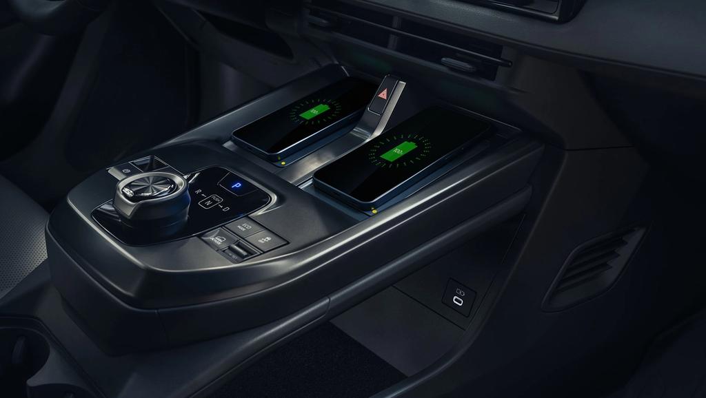 Close-up of a car’s center console featuring two wireless charging pads with glowing green indicators, a rotary control dial with buttons, a gear selector, and a USB port below. The interior is modern and sleek.
