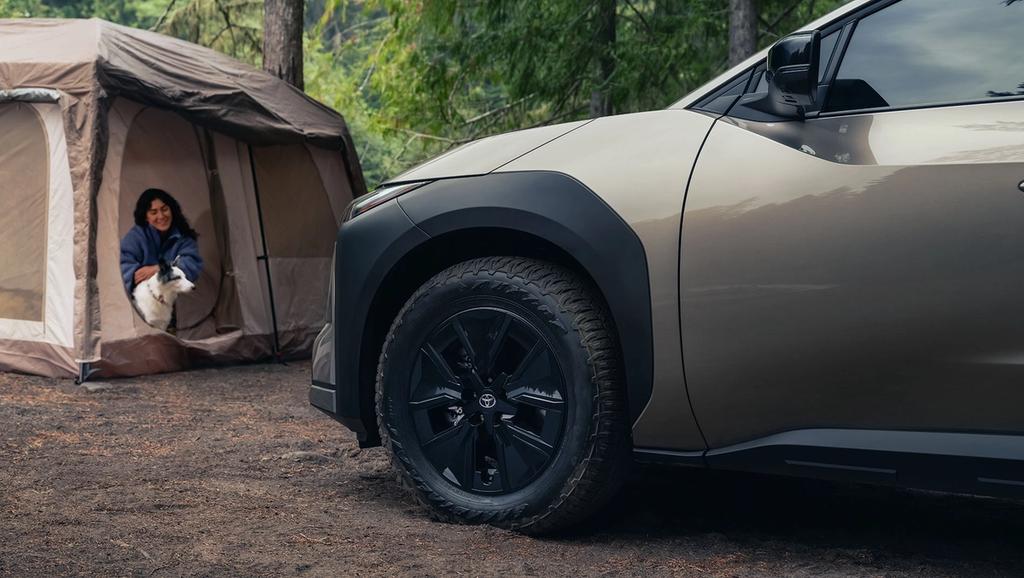 A woman sits at the entrance of a camping tent in a forest, petting a dog, while the front side of an SUV is parked nearby on the dirt ground.