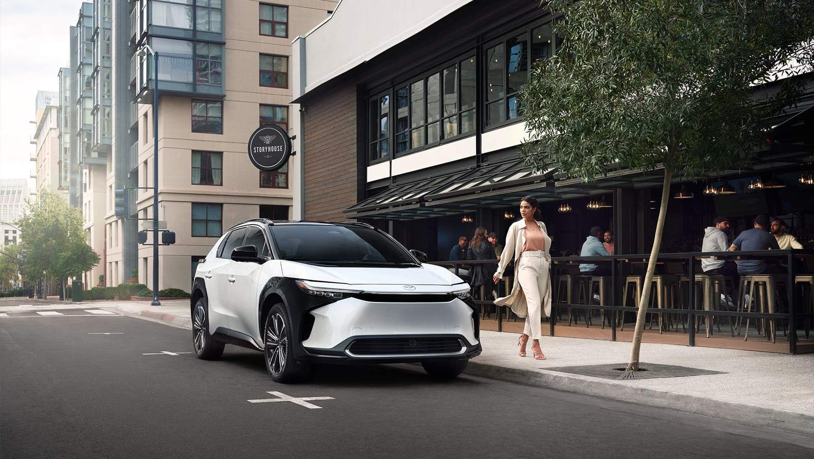 2024 white Toyota bZ4x all electric plug in vehicle parked on a city street in front of a restaurant with people eating on the patio