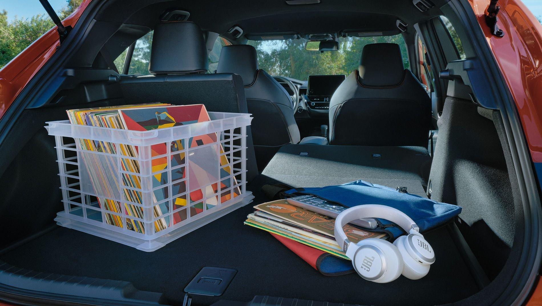 A car trunk with folded rear seats holds a crate of vinyl records, a stack of magazines, a pair of white JBL headphones, and a blue bag
