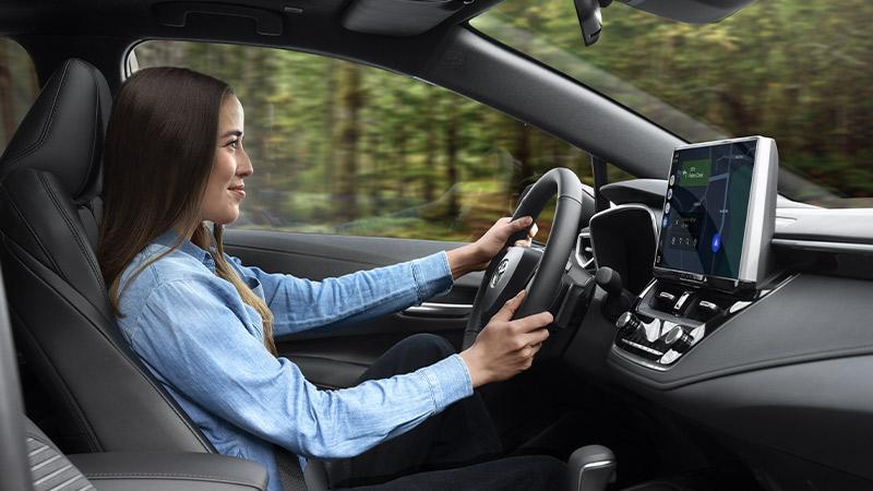 A woman wearing a light blue shirt smiles while driving a car through a forested area, with a navigation screen visible on the dashboard.