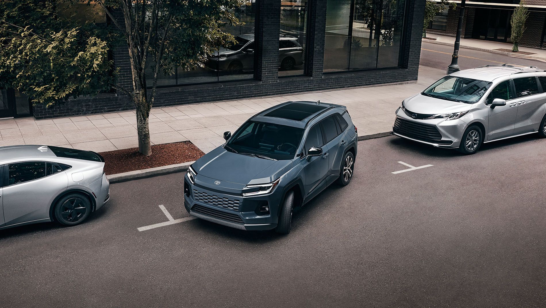 A blue SUV is parallel parked between a silver sedan and a silver minivan on a city street next to a sidewalk with trees and a brick building.