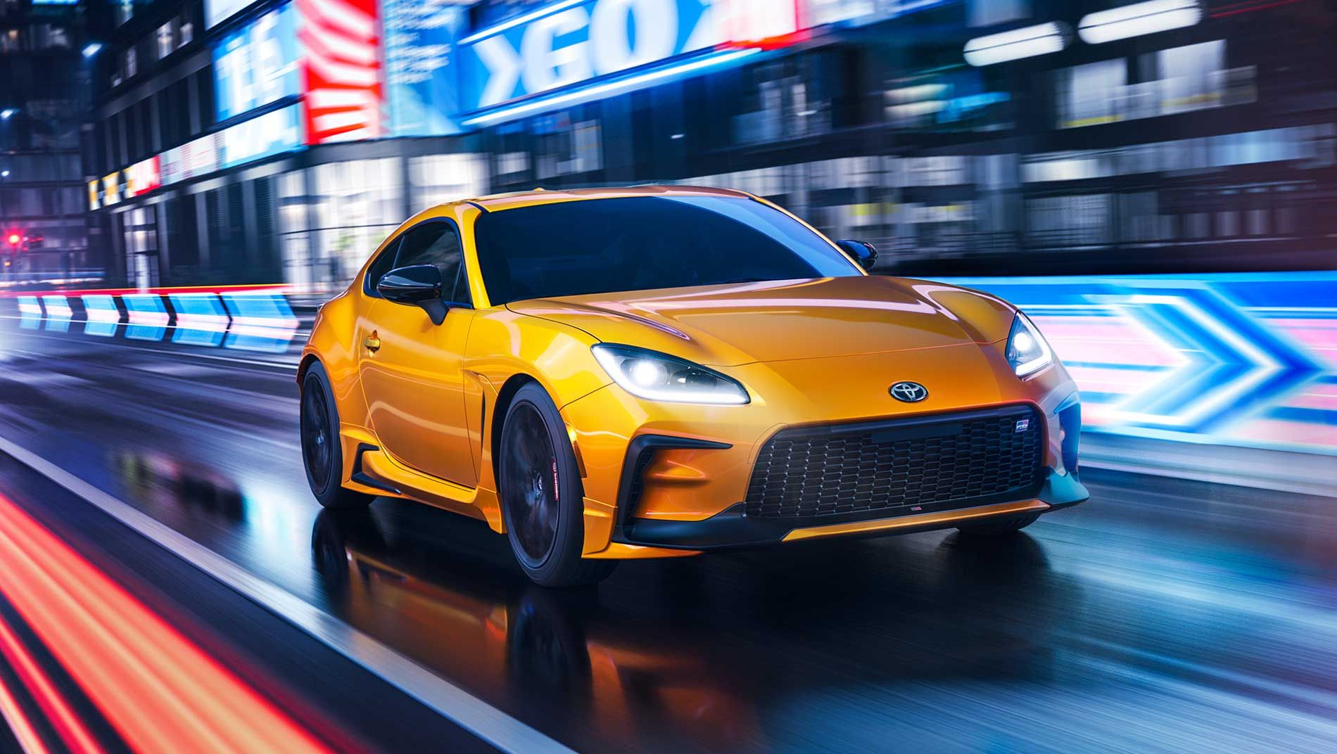 A bright yellow toyota gr86 drives quickly through a city street at night, with blurred neon lights