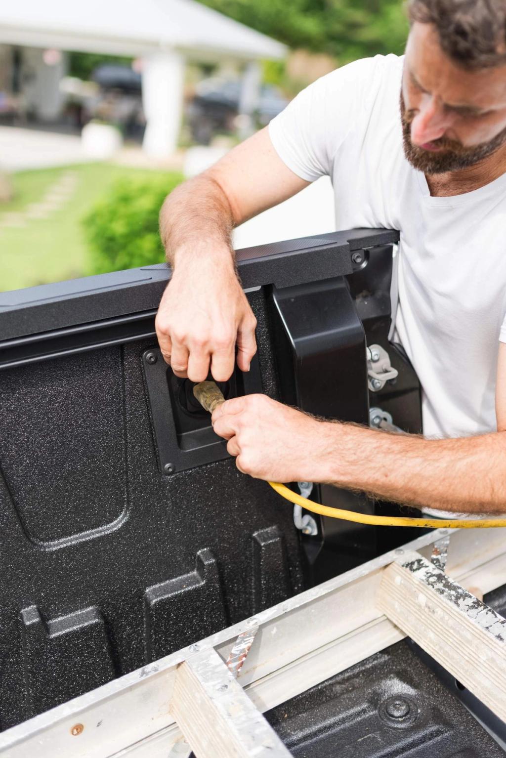 A man plugs a yellow electrical cord into a power outlet located on the inside wall of a pickup truck bed.