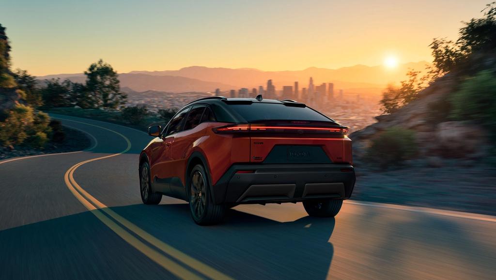A red electric SUV drives on a winding road through hills at sunset, with a city skyline and mountains visible in the distance under a clear sky.