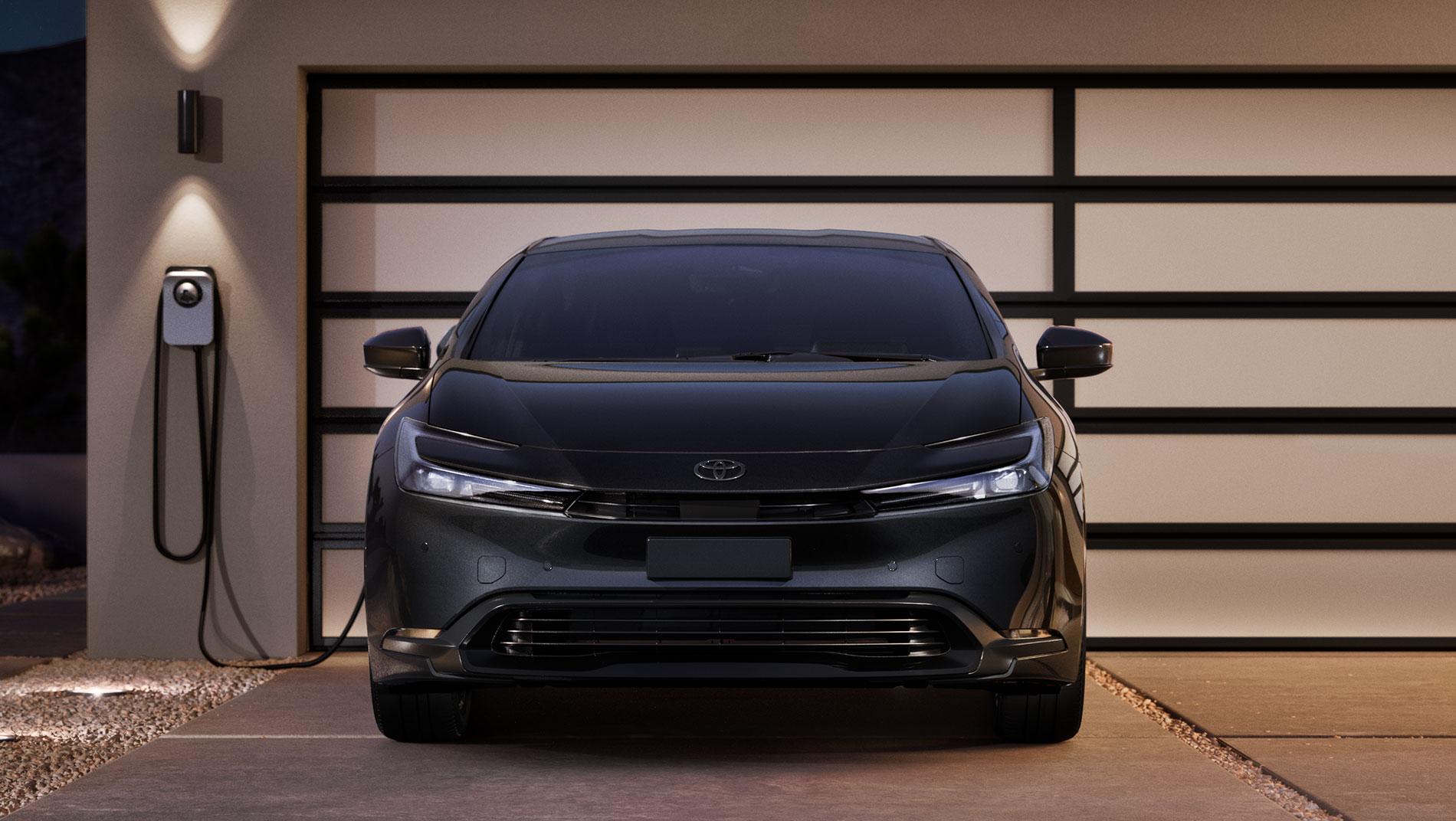 A black Toyota 2025 prius plug in is parked in front of a modern garage door, connected to an electric vehicle charging station mounted on the wall.