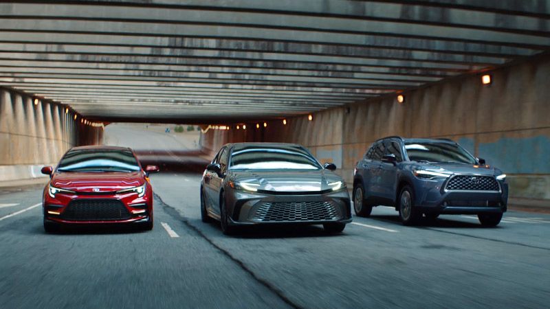Three cars—a red sedan, a silver sedan, and a blue SUV—drive side by side through a wide, well-lit tunnel on a multi-lane road. The tunnel walls are lined with concrete and the scene appears dynamic.