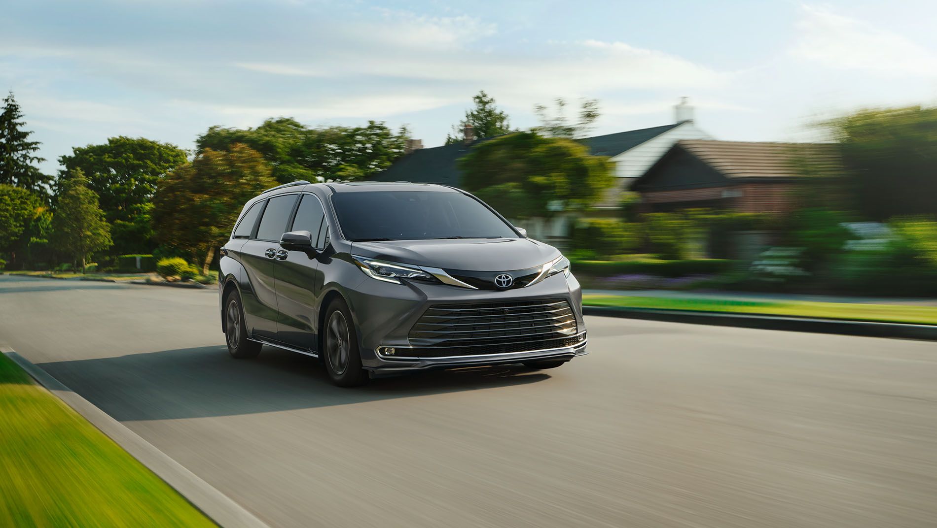 A dark gray Toyota minivan is driving on a suburban street with green lawns, trees, and houses in the background on a sunny day. The image has a sense of motion blur.