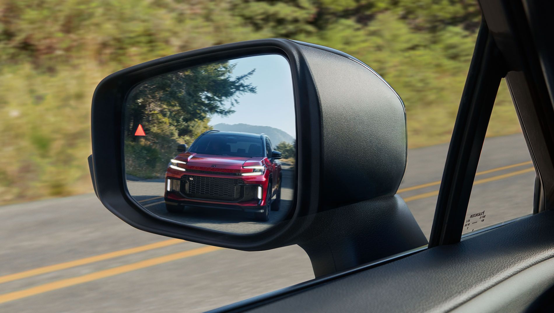 A red SUV is visible in the side mirror of a car driving on a two-lane road, surrounded by trees and greenery under a clear sky.