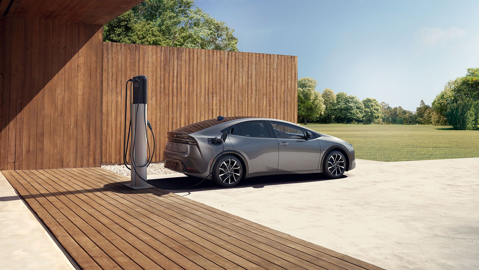 A silver electric car is parked next to a wooden building, charging at an outdoor charging station on a sunny day, with green trees and grass in the background.