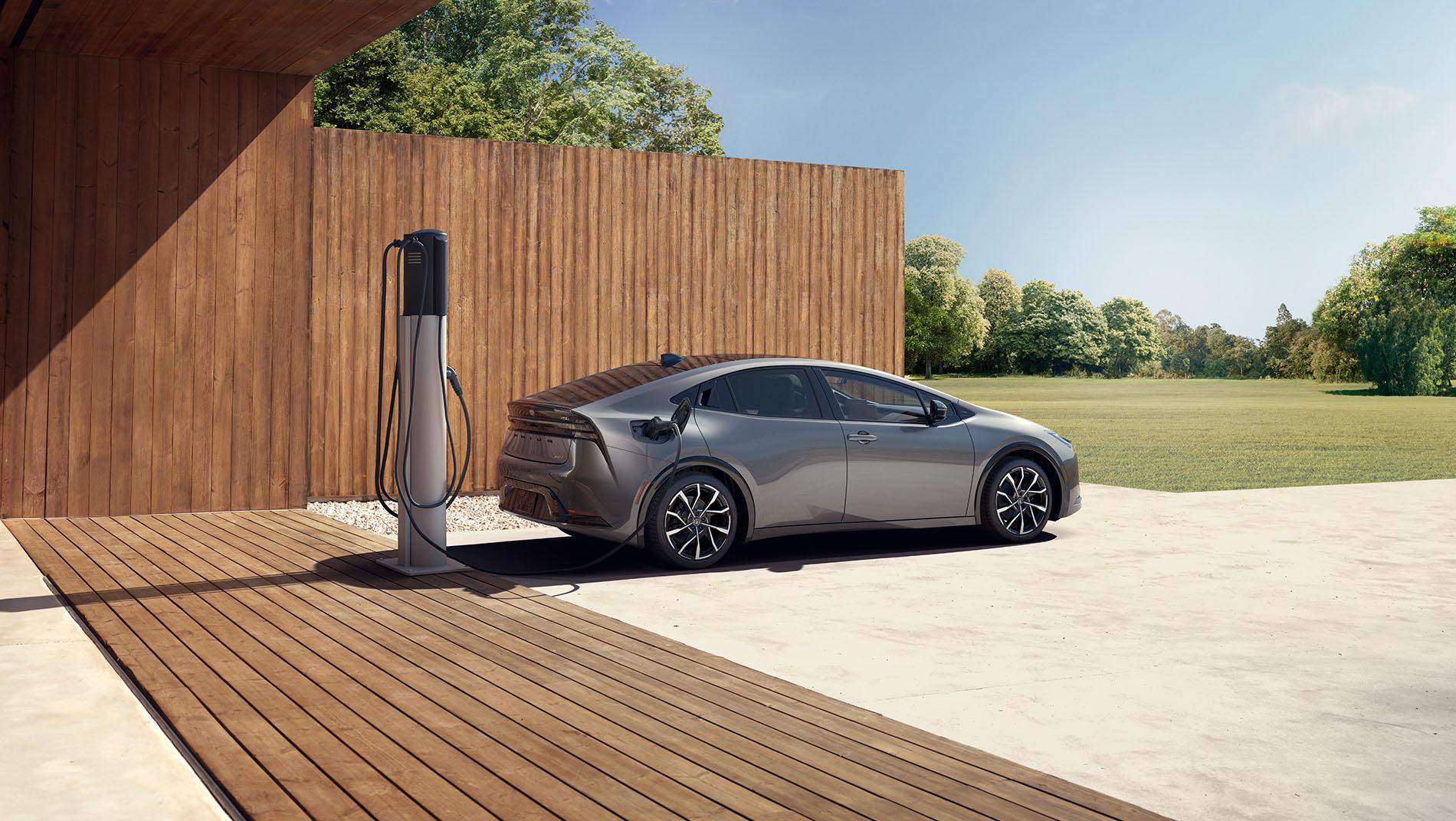 A silver electric car is parked next to a wooden building, charging at an outdoor charging station on a sunny day, with green trees and grass in the background.