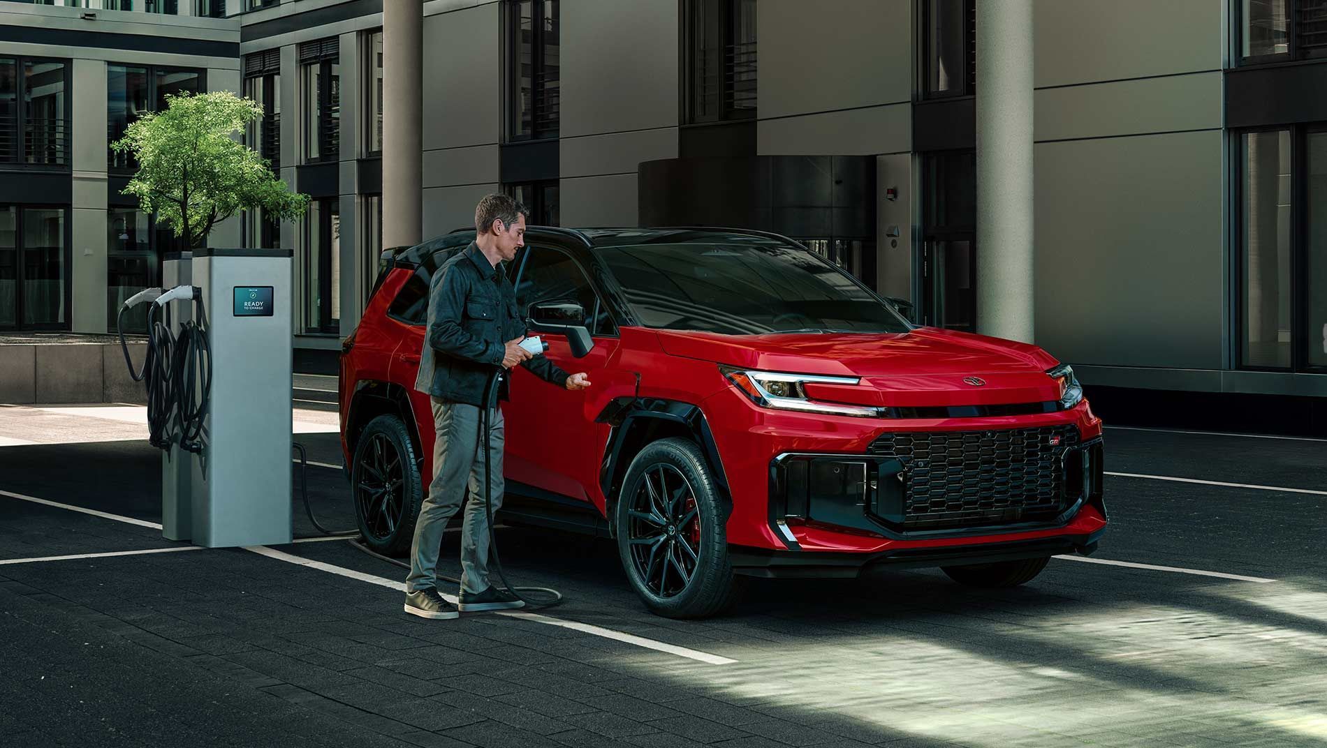 A man stands next to a red electric RAV4 Plugin SUV, plugging a charging cable into the vehicle at a charging station