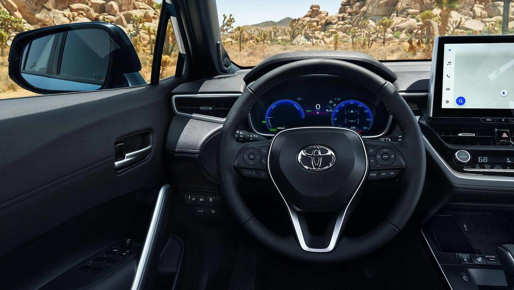 View of a Toyota car interior from the driver's seat, showing the steering wheel with Toyota logo, digital dashboard, infotainment screen with a map, and desert landscape visible through the window.