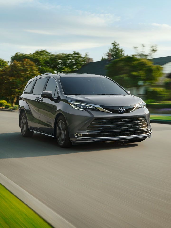 A dark gray Toyota minivan drives down a suburban street with green trees, lawns, and houses in the background under a partly cloudy sky.