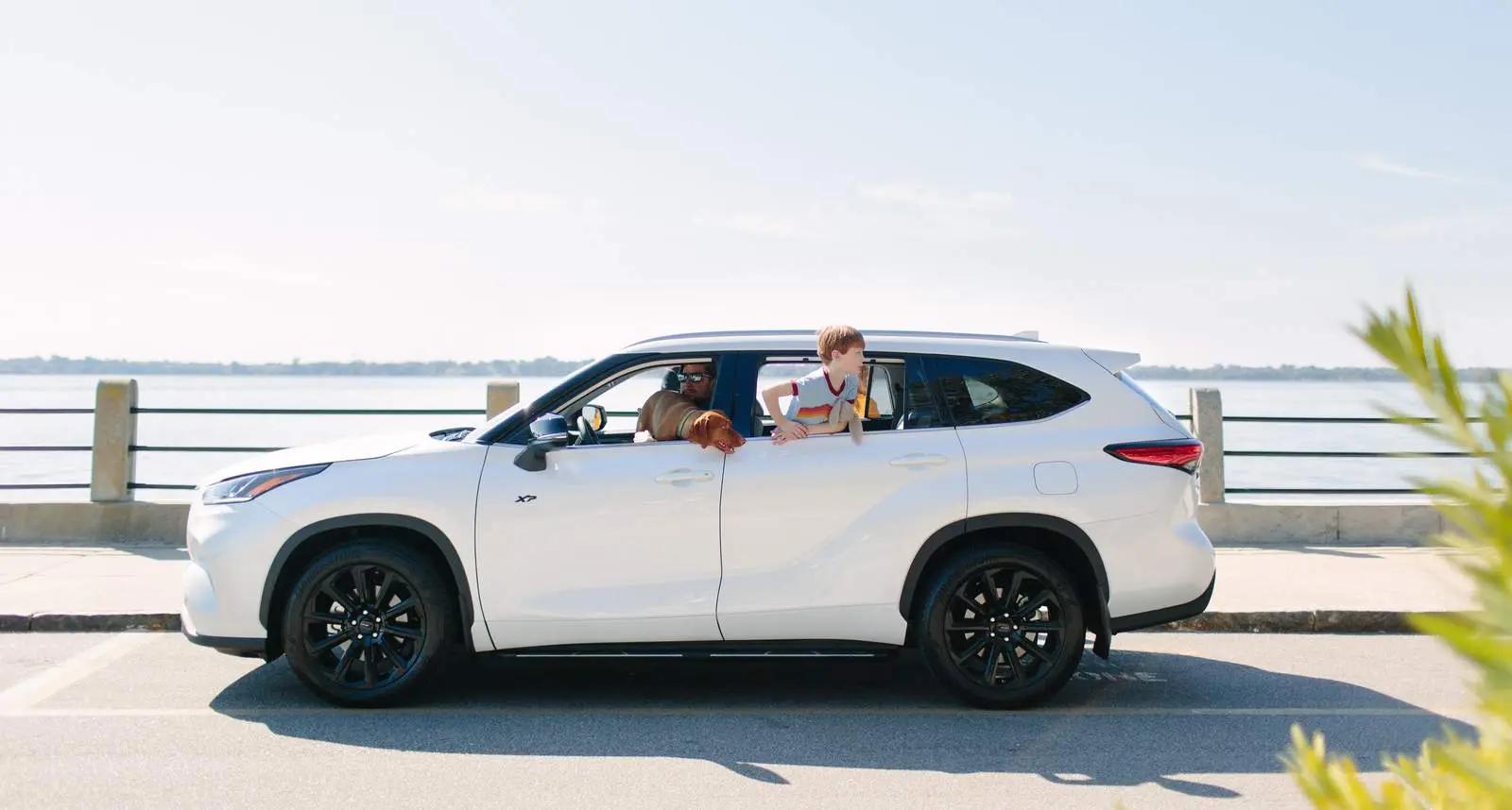 A white xseries SUV is parked near a waterfront. A dog and a young boy are looking out of the back window, while a person sits in the driver's seat.