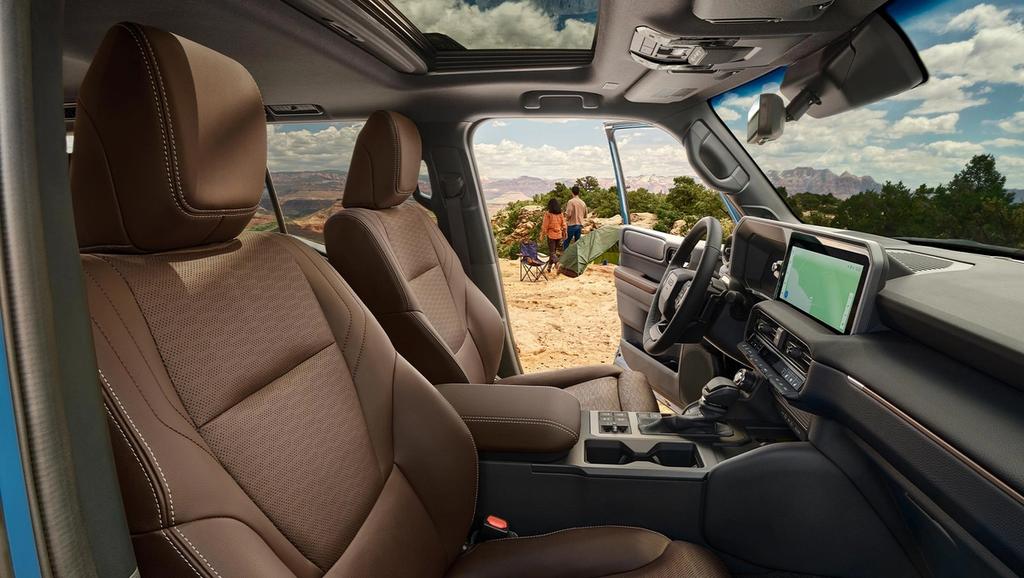 inside the front of the 2025 Toyota Land Cruiser hybrid with the sunroof and navigation system. the drivers door is open showing 2 people setting up camp on a mountain top