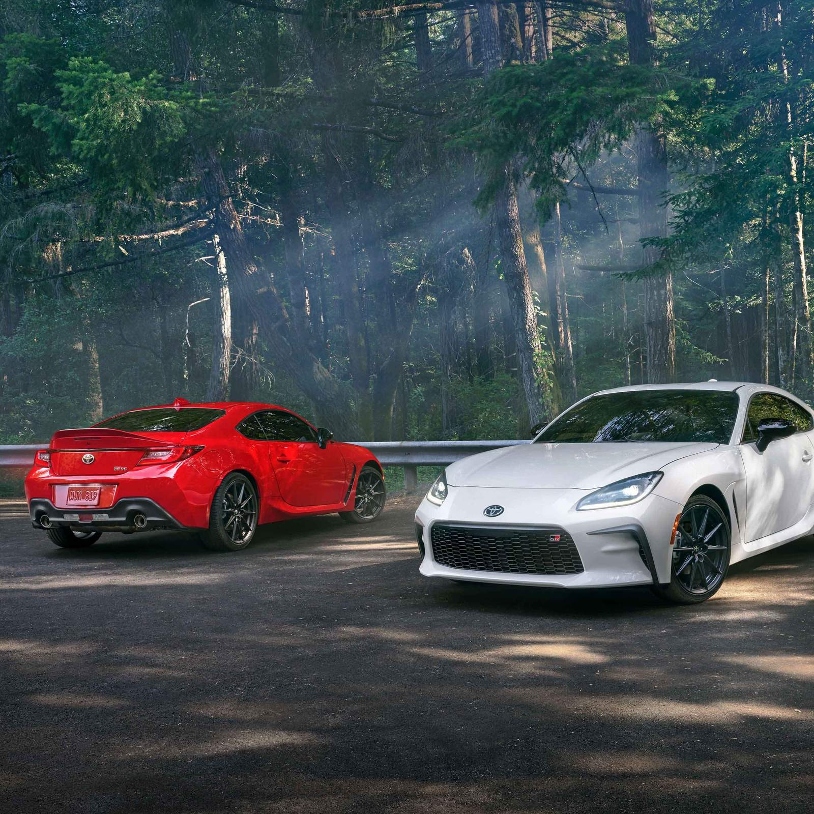 2 Toyota GR86 - one white one red parked in a parking lot in a forest of trees