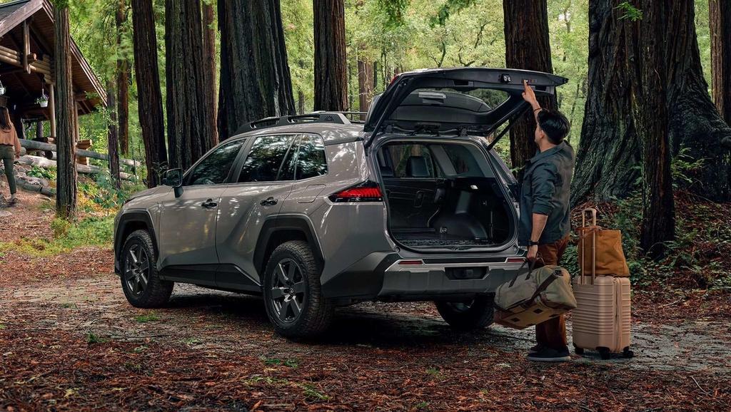 A person stands next to an SUV with its trunk open, holding luggage, surrounded by tall trees in a forest near a wooden cabin.