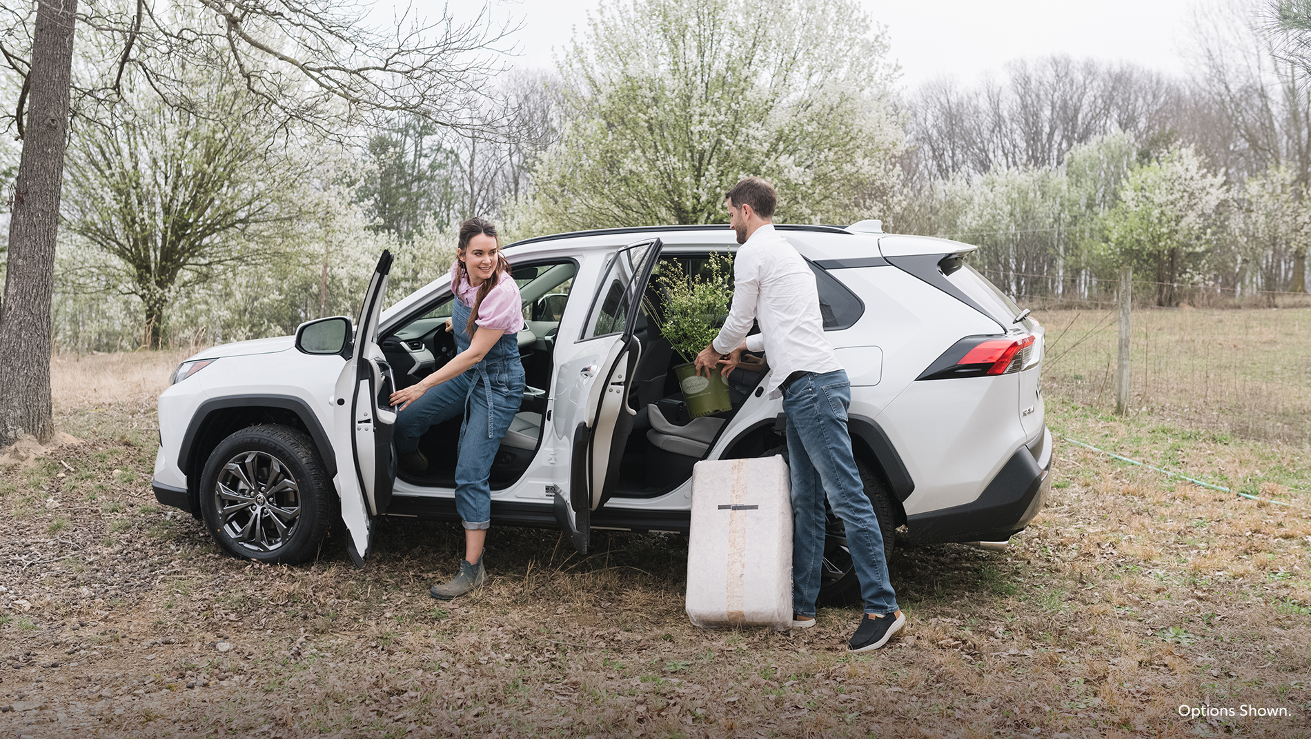 a White RAV4 Hybrid beyond zero vehicle parked in a field with a man and woman getting plants out of the backseat