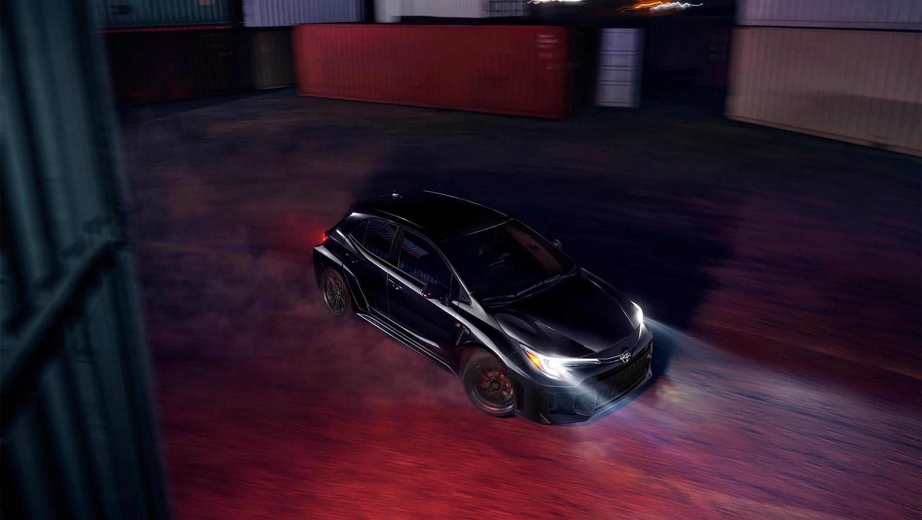 A black 2025 toyota gr corolla drifts in an industrial area with containers in the background. The car's headlights illuminate the ground.