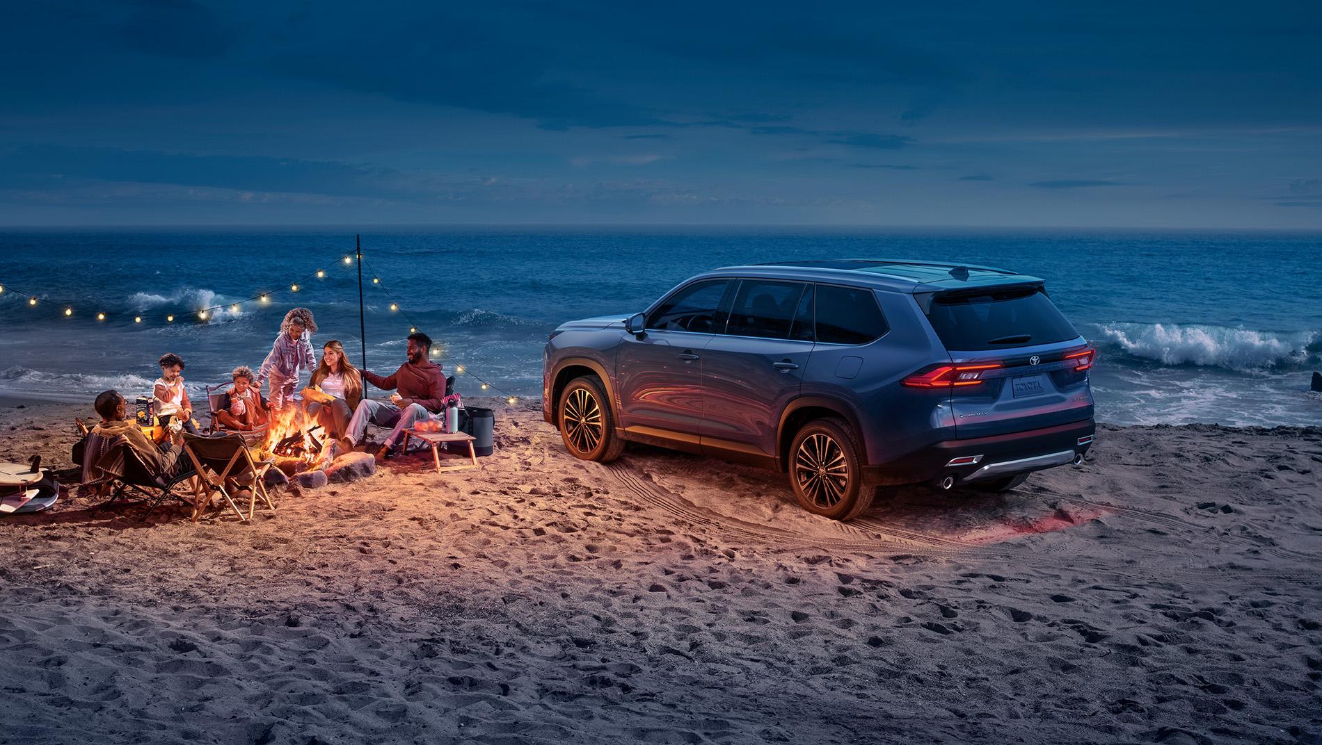 2024 Toyota Grand Highlander Hybrid parked on a beach while the family enjoys a bonfire on the beach at sunset