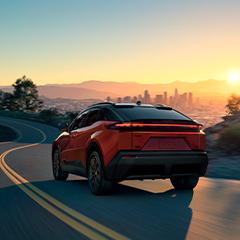 A red electric car drives on a winding road toward a city skyline at sunset, with mountains in the background and trees lining the roadside.