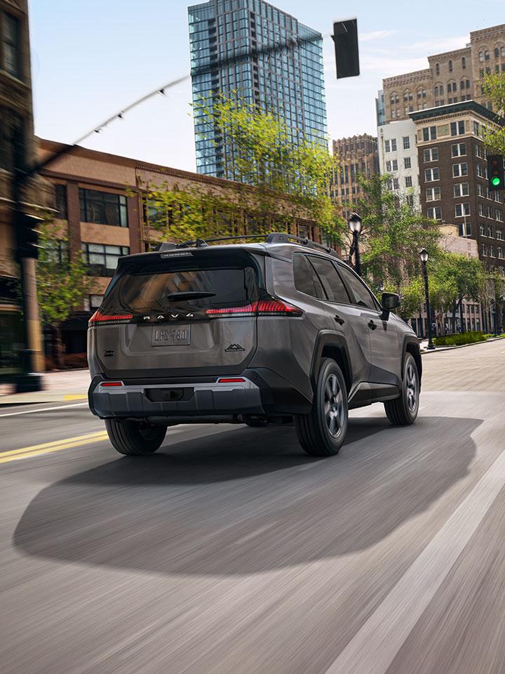 A dark gray SUV drives down a city street lined with modern and historic buildings, passing under a traffic light on a sunny day. Trees and tall glass skyscrapers are visible in the background.