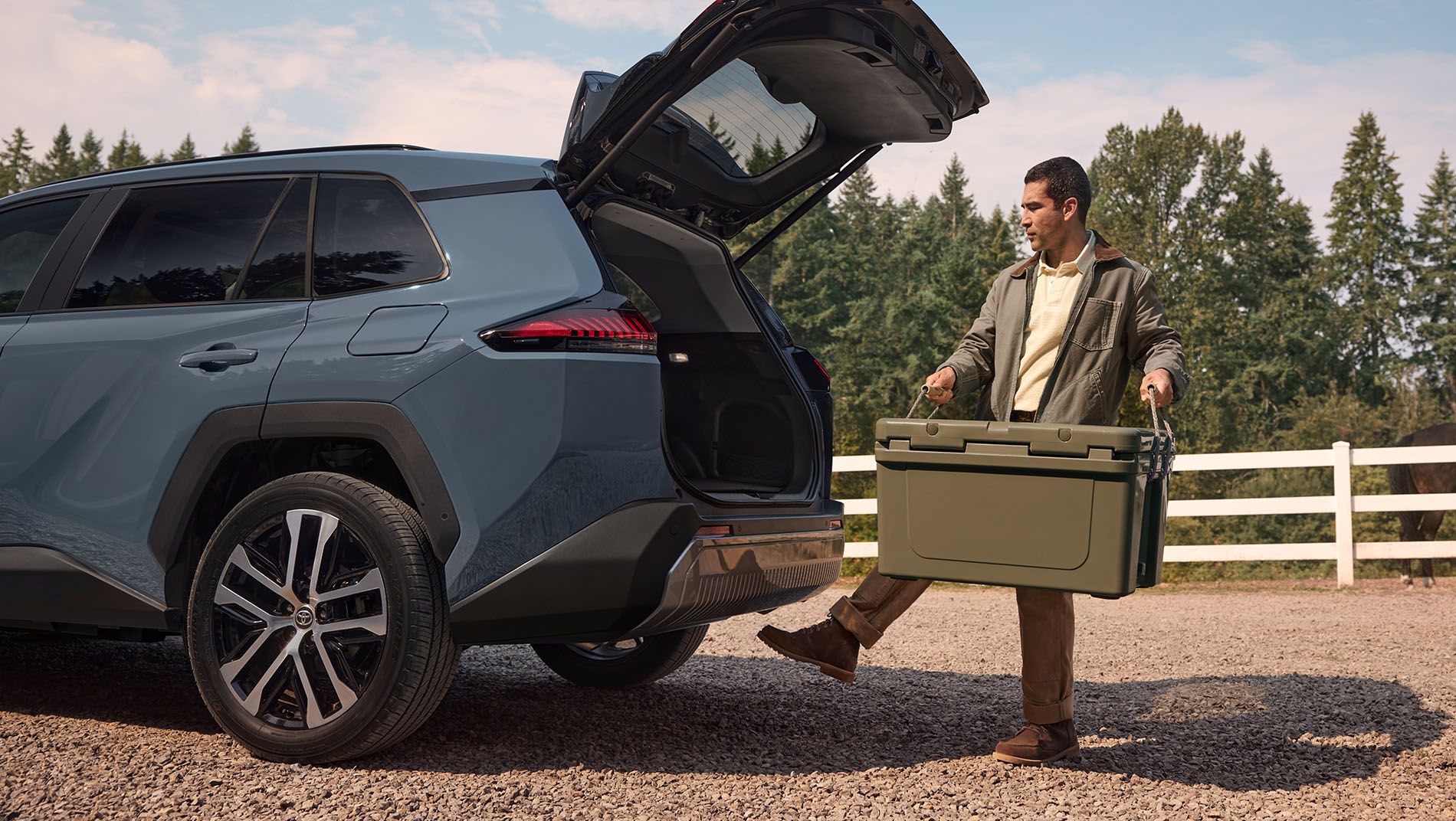 A man stands behind an SUV with the trunk open, holding a large green cooler and preparing to load it. Trees and a white fence are visible in the background on a sunny day.