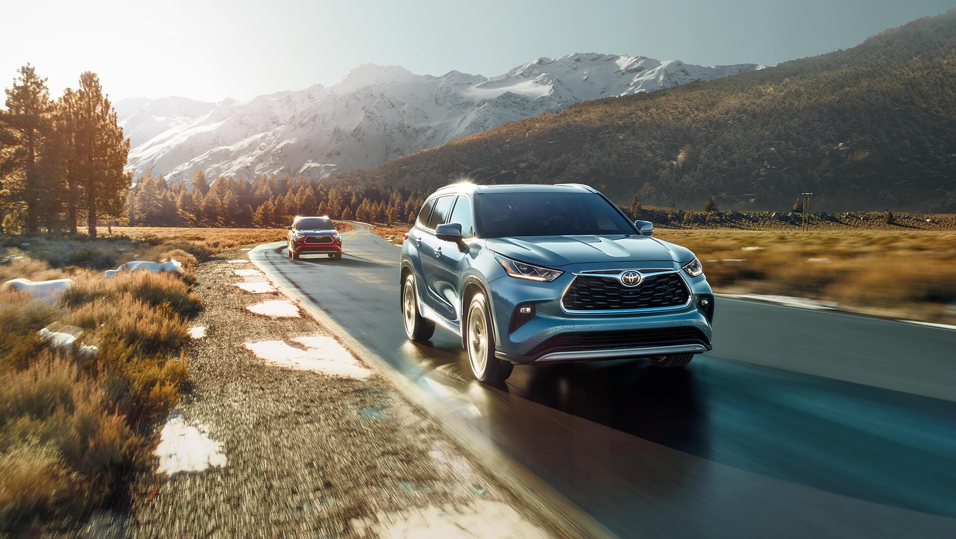 blue Toyota Highlander SUV being followed by a red Highlander driving down a road with mountains behind