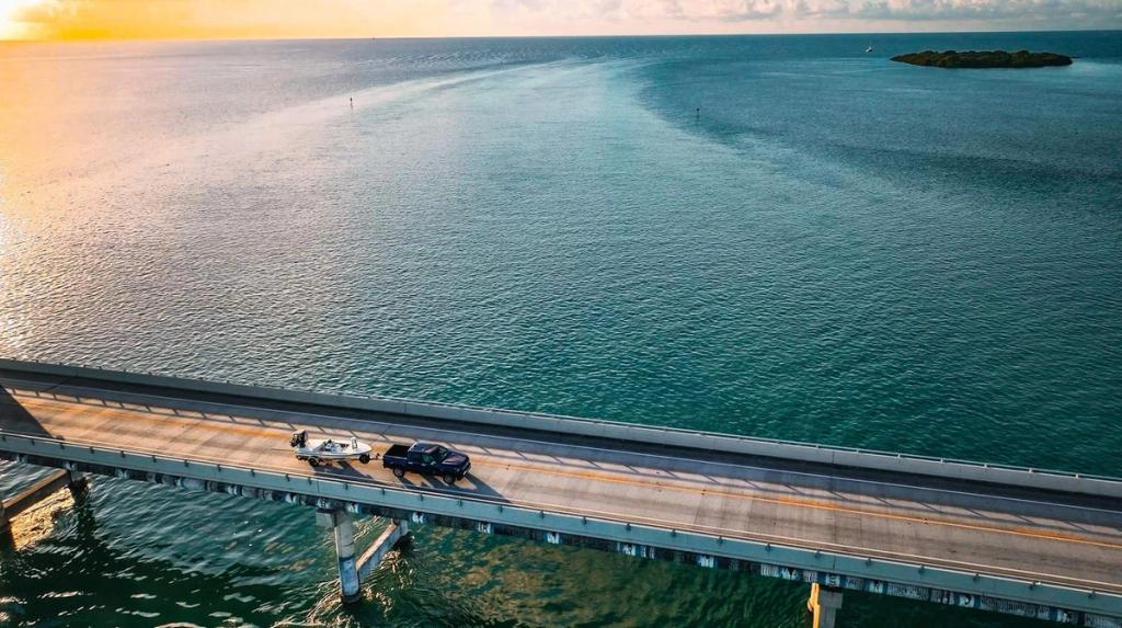 A car towing a boat travels across a long bridge over calm blue-green water at sunset, with an island visible in the distance and the sky tinged with orange and yellow hues.