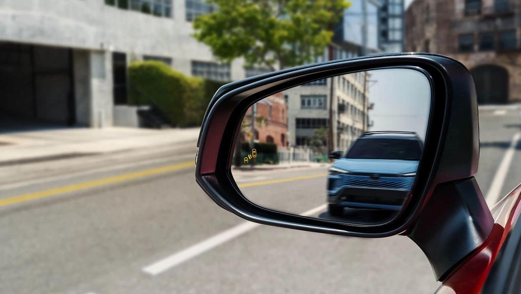 A close-up of a car's side mirror shows another vehicle approaching from behind on a city street. The mirror displays a yellow blind spot warning indicator.