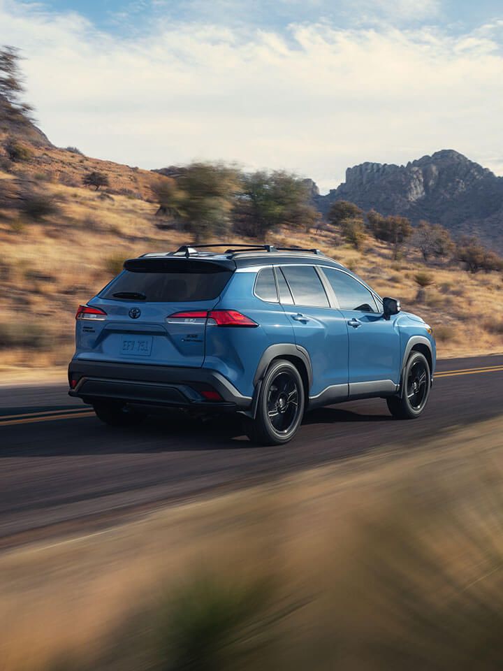 A blue Corolla Cross SUV drives along a scenic, winding road bordered by dry grass and rocky hills