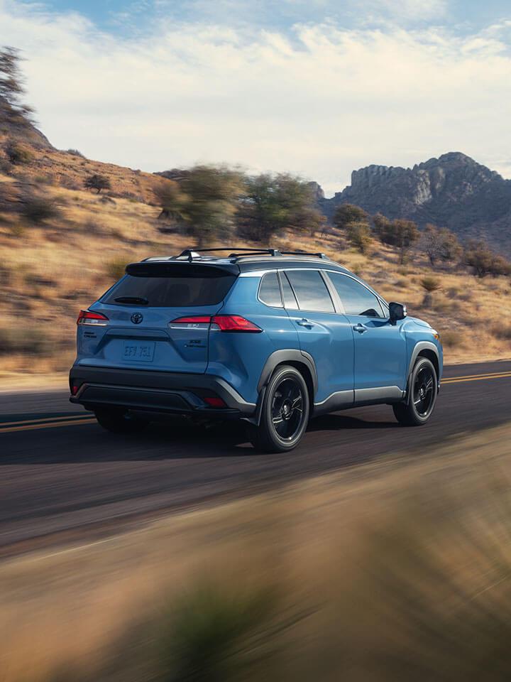 A blue Corolla Cross SUV drives along a scenic, winding road bordered by dry grass and rocky hills