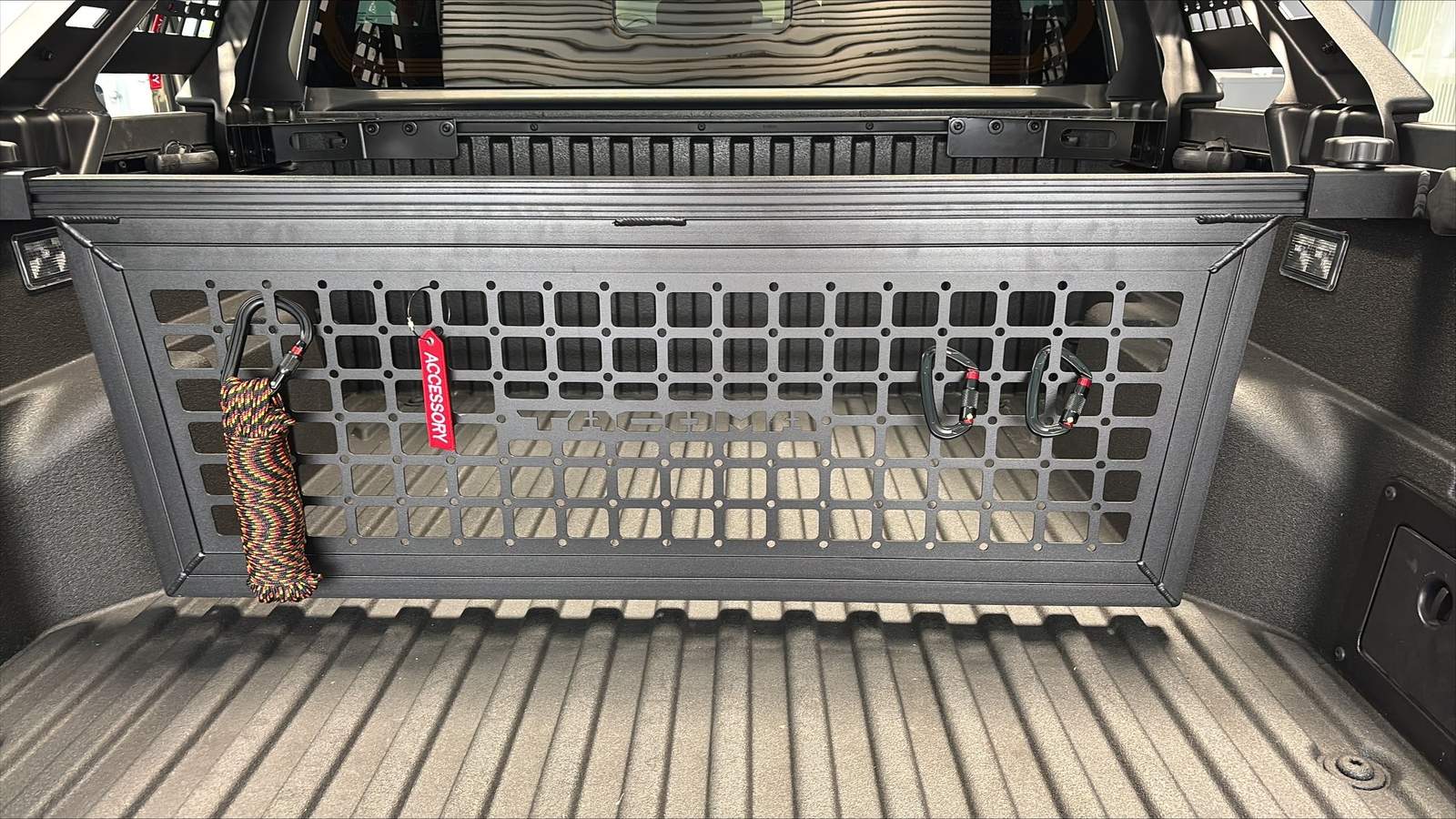 The image shows the bed of a toyota pickup truck with a modular grid panel attached to the back. The panel has a black design with attachment points.