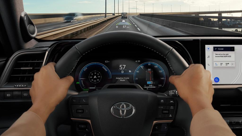 image from the drivers perspective as they hold the wheel of a 2025 Toyota Crown Hybrid and see the large dashboard, multimedia touch screen, and head up display in the windshield