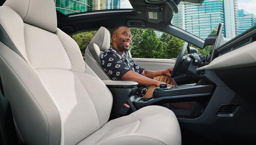 A man smiles while sitting in the driver's seat of a modern car with a light-colored interior; city skyscrapers and greenery are visible through the windows.