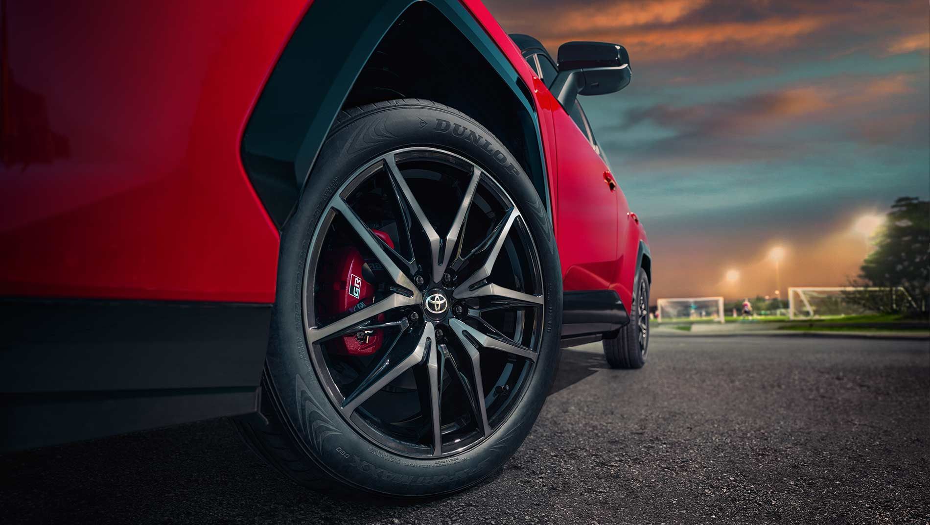 Close-up view of a black alloy wheel and tire on a red rav4 vehicle, parked on asphalt near a sports field at sunset