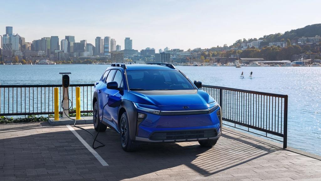 A blue electric SUV is parked at a charging station beside a waterfront, with a city skyline and a few paddle boarders visible in the background on a sunny day.
