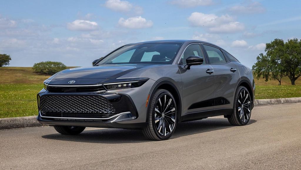 A sleek, gray modern sedan with black accents parked on a paved road, surrounded by grass, trees, and a partly cloudy sky in the background.