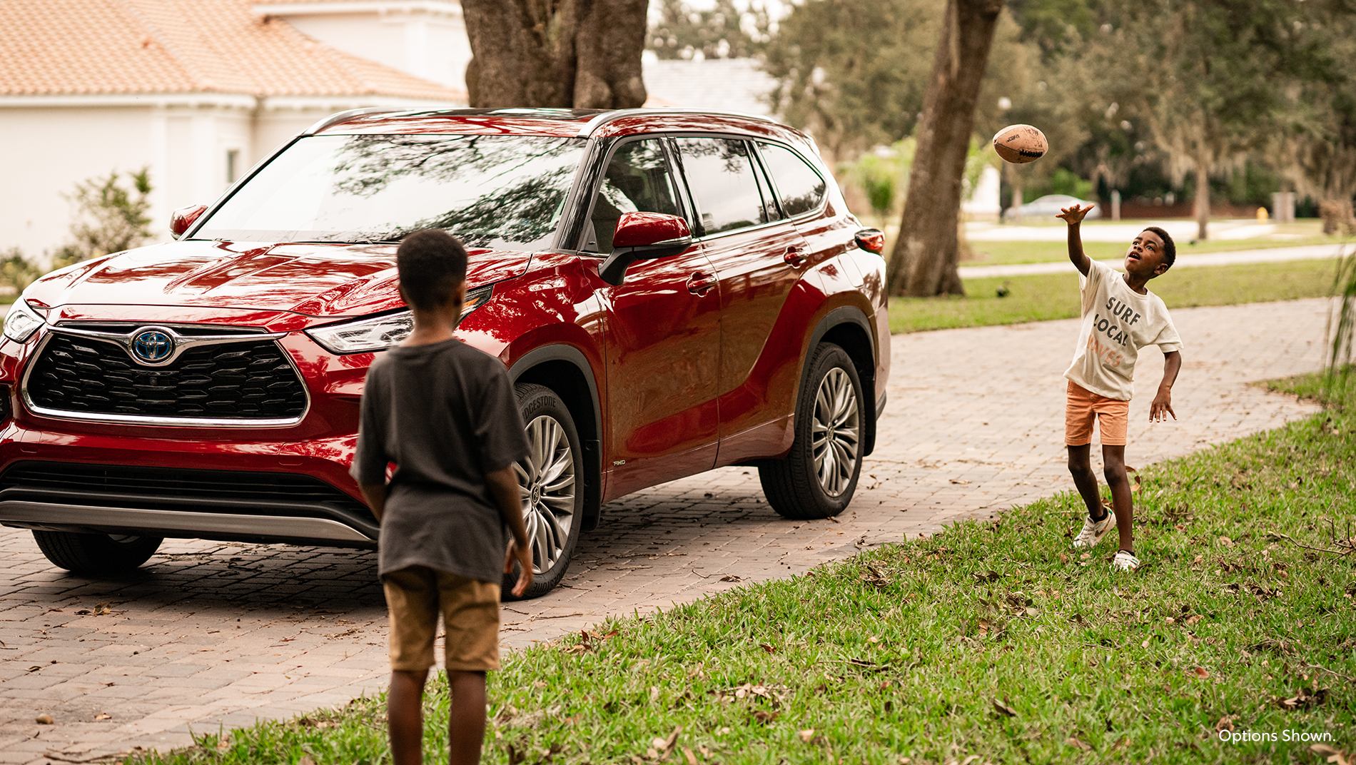 a Red Toyota Hybrid SUV is parked in a suburban driveway while 2 boys play football in the front yard