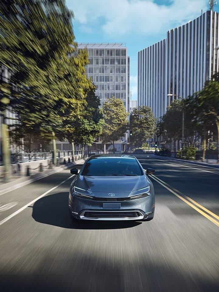 Front shot of a 2024 Grey Toyota Prius Prime Plug in hybrid driving down a city street covered by the Toyotacare Toyota maintenance plan