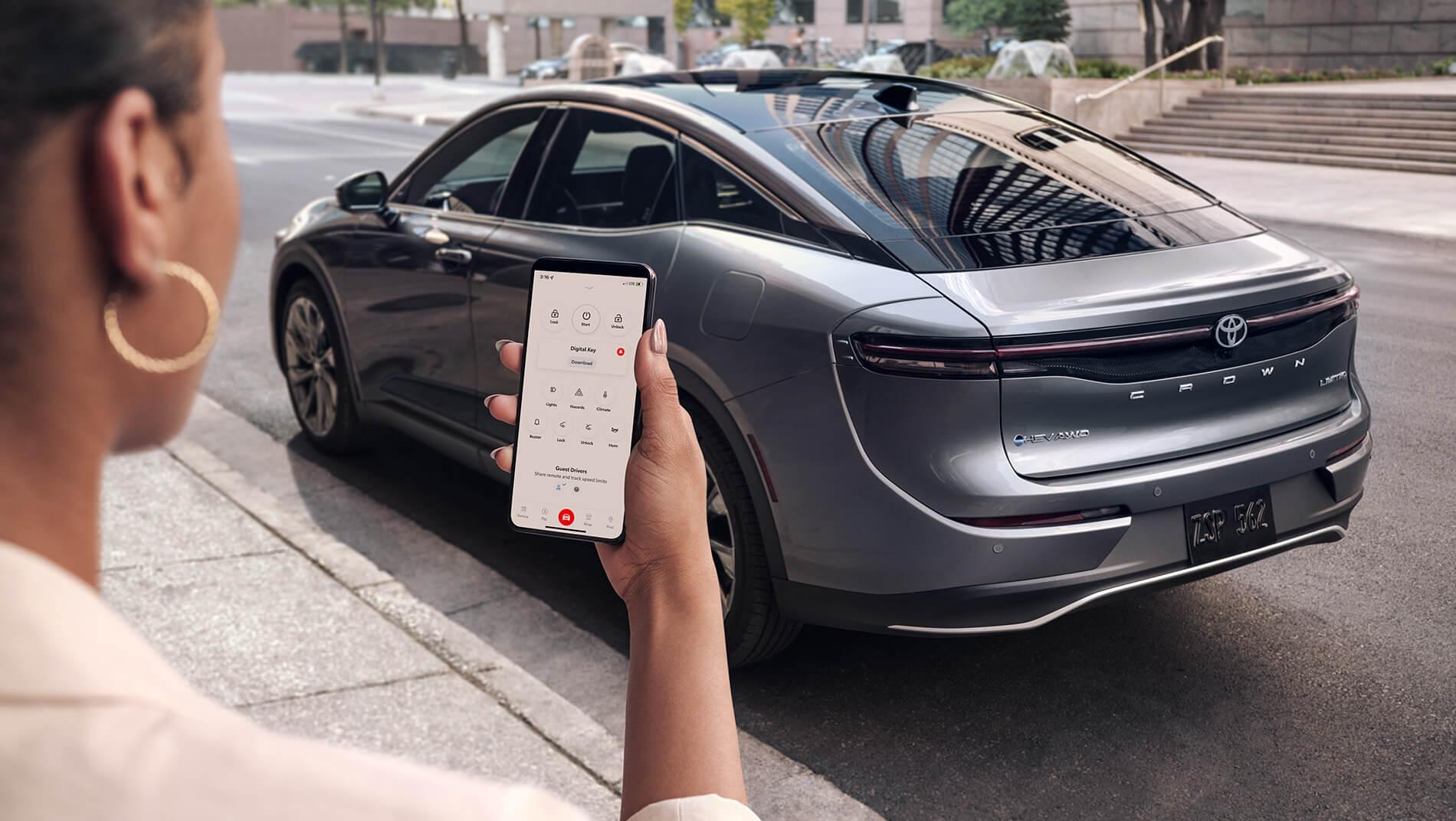 Woman on city street using her Toyota Connect App on her smart phone to unlock and start the grey Toyota Crown sedan parked at the curb