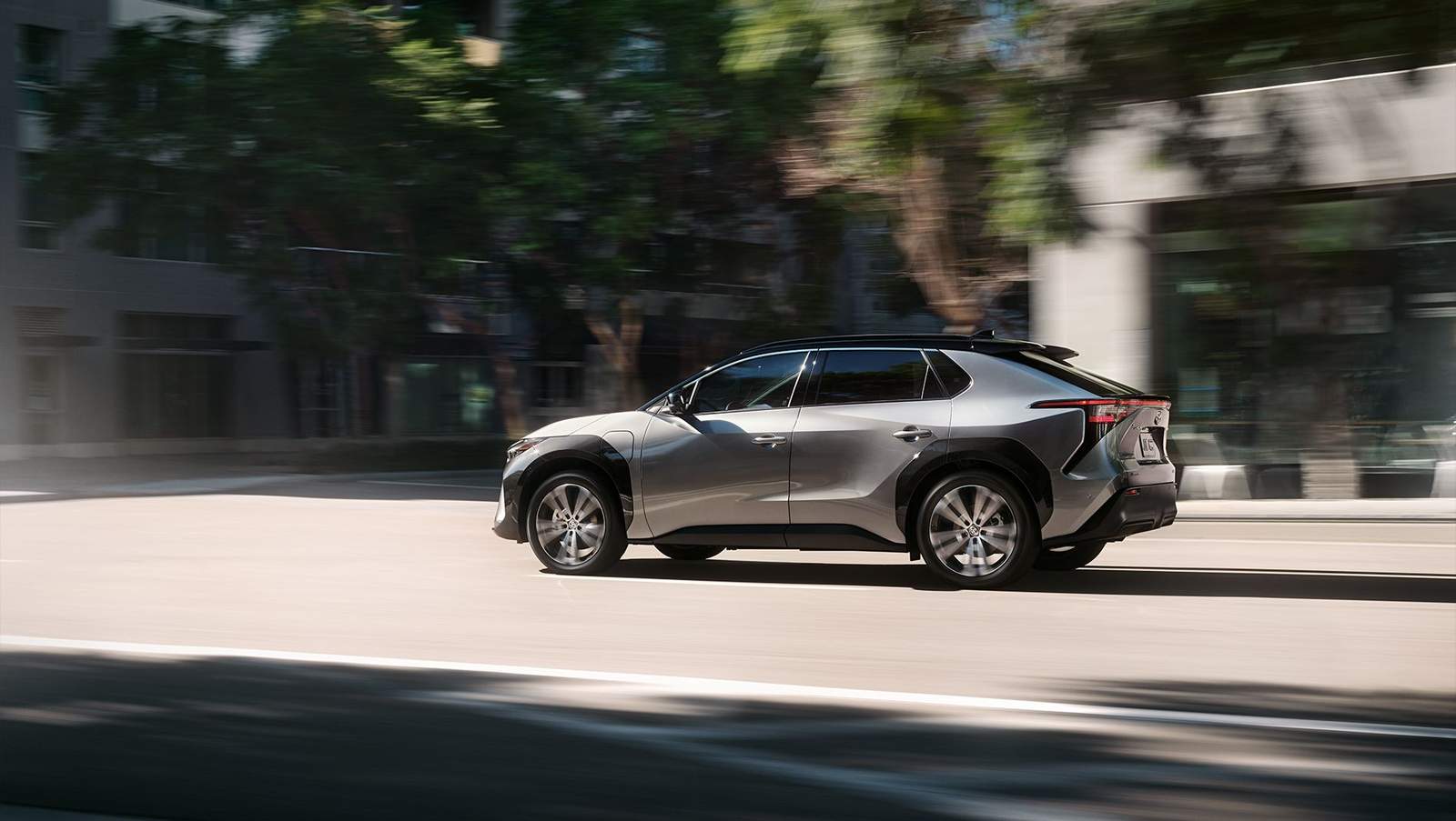2024 silver Toyota bZ4x all electric plug in vehicle driving down a tree lined city street