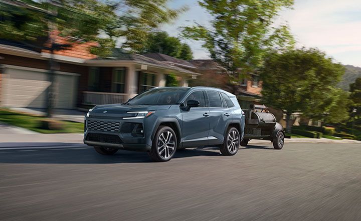 A gray SUV drives down a suburban street, towing a small black trailer smoker behind it. Houses with porches and green lawns line the background, with trees and blurred motion suggesting speed.