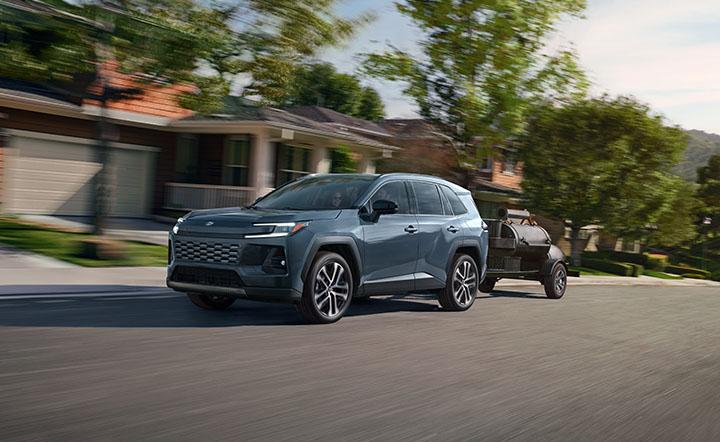 A gray SUV drives down a suburban street, towing a small black trailer smoker behind it. Houses with porches and green lawns line the background, with trees and blurred motion suggesting speed.