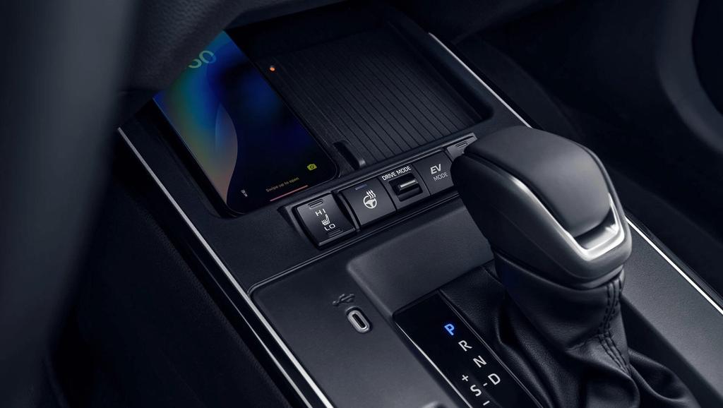 Close-up of a car’s center console showing a gear shifter, electronic parking brake and drive mode buttons, and a smartphone partially placed in a compartment above the controls.