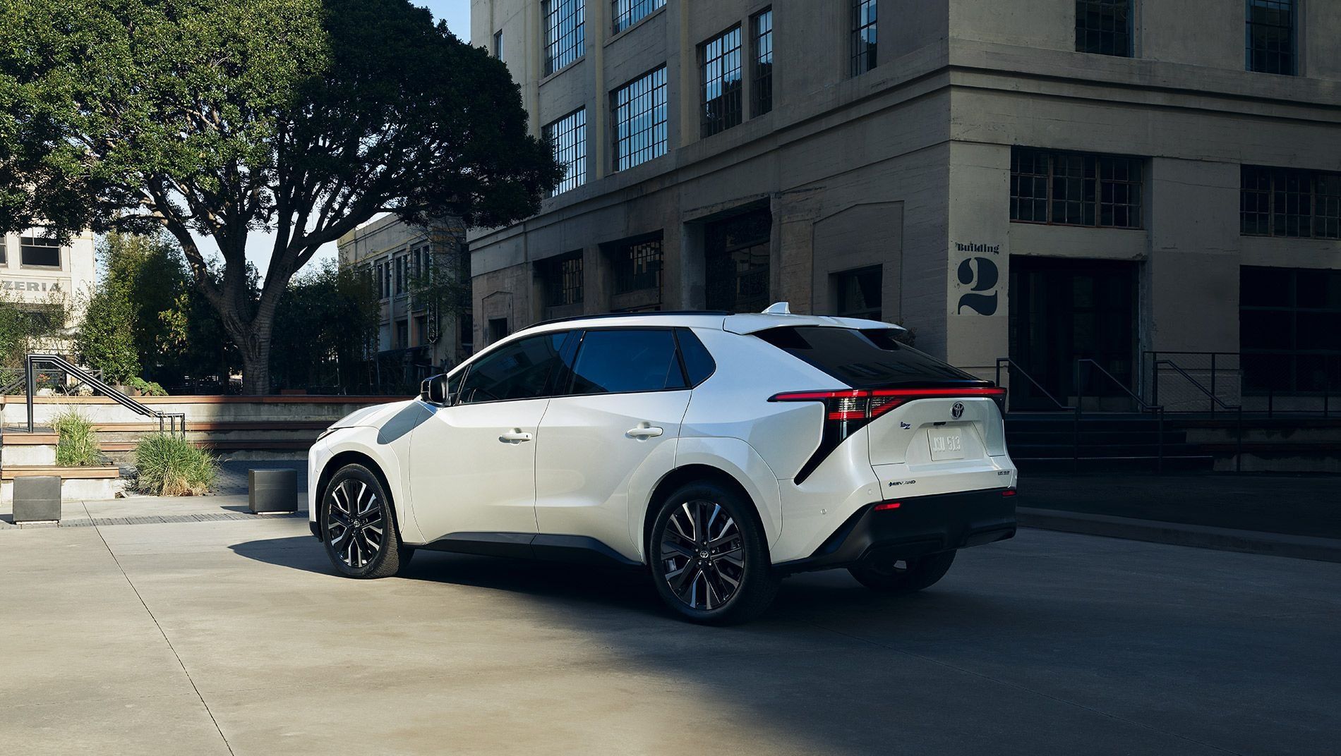 A white toyota bZ is parked on a sunlit street in front of a modern industrial-style building with large windows, trees, and steps in the background.