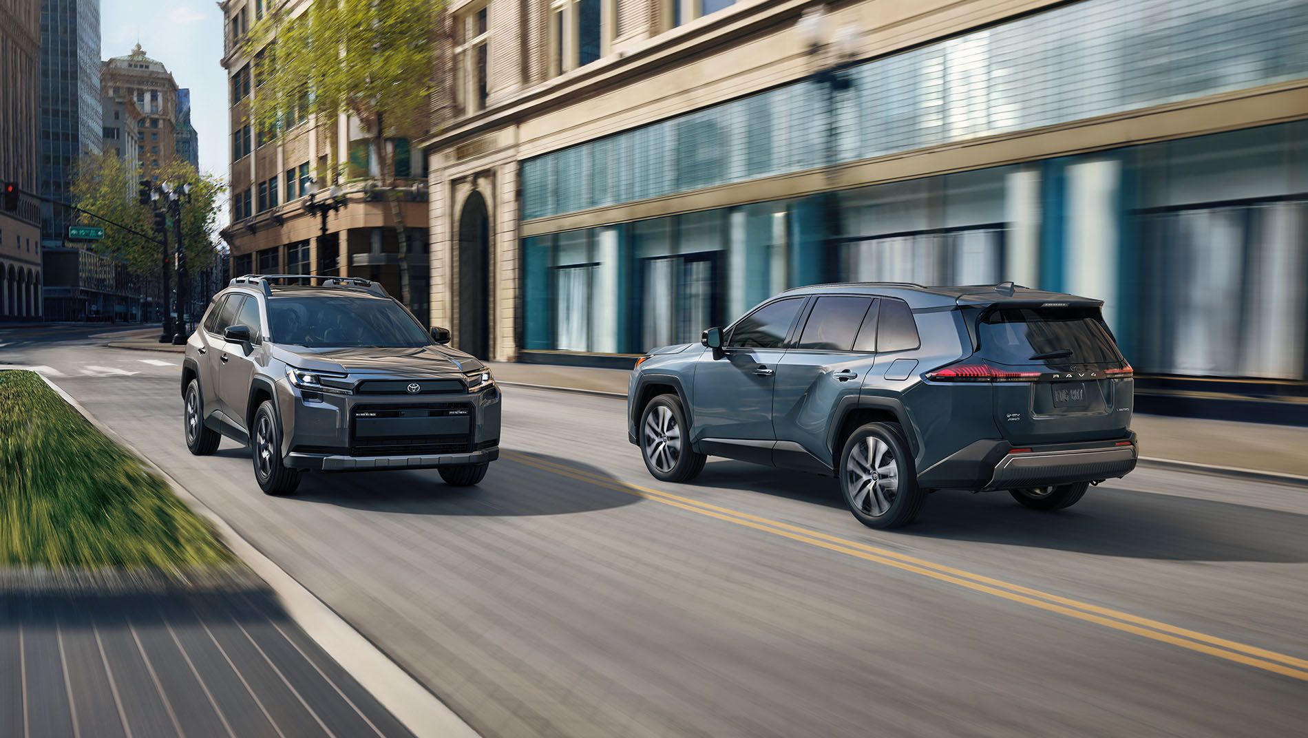 Two modern SUVs drive side by side on a city street lined with tall buildings and trees, with blurred motion indicating speed. Both vehicles appear to be Toyota RAV4 models in different colors.